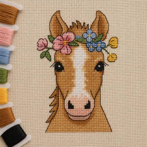 May include: A cross-stitch embroidery of a brown horse head with a white blaze on its face, wearing a floral crown of pink, blue, and yellow flowers. The design is stitched on beige Aida fabric, accompanied by a vertical row of colourful embroidery floss.