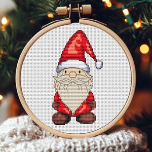May include: A cross-stitch embroidery of a Santa Claus gnome in a wooden hoop. The gnome wears a red hat and coat, a white beard, and brown boots. The background is blurred with Christmas tree branches and lights.