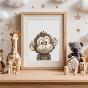 Op de afbeelding: Een ingelijst kruissteekkunstwerk met een lachende cartoon-aap met bruine vacht en grote oren. Het kunstwerk staat in een licht houten frame, tentoongesteld op een houten oppervlak met speelgoeddieren en decoratieve sterren.