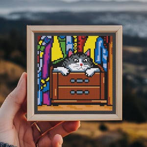 May include: Cross-stitch art of a gray and white cat peeking out of a brown dresser. The cat is in a room with colorful clothing. The artwork is framed in a light wood frame.