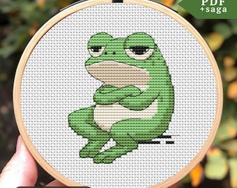 May include: A cross-stitch pattern of a green frog with its arms crossed, sitting on a white background. The pattern is in a wooden embroidery hoop. The text "Stitches: 40 x 47" is below the hoop. The text "PDF + saga" is in the upper right corner of the image.