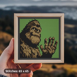 Op de afbeelding: Kruissteekkunst van een Bigfoot-wezen, omlijst in licht hout, tegen een groene achtergrond. De Bigfoot is weergegeven in bruin en zwart. De tekst "Stitches: 65 x 65" staat onderaan. Afmetingen: 165 cm x 165 cm.