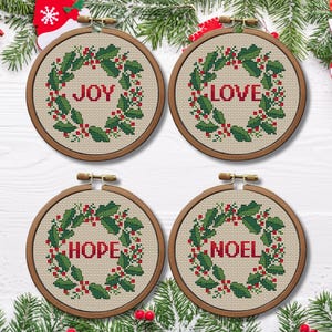 May include: Four circular cross-stitch hoops with festive messages. Each hoop displays a wreath of green leaves and red berries encircling the words "JOY", "LOVE", "HOPE", and "NOEL" in red. The hoops are set against a backdrop of evergreen branches.