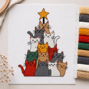 May include: A cross-stitch pattern on white Aida fabric featuring a pyramid of cats in various colors including red, gray, white, orange, and green, topped with a yellow star. A set of embroidery floss bobbins is aligned on the right side.