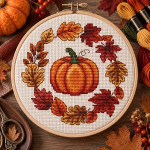 Puede incluir: Un bastidor de bordado circular de madera que contiene un diseño de punto de cruz de una calabaza naranja central rodeada por una corona de hojas de otoño coloridas en tonos rojos, naranjas y amarillos sobre un fondo de tela blanca.