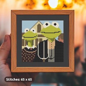 Op de afbeelding: Kruissteekkunstwerk in een houten lijst. De afbeelding toont twee groene kikkerpersonages in formele kleding, voor een gebouw. De achtergrond is een lichtblauwe lucht. De tekst "Stitches: 65 x 65" staat onderaan.
