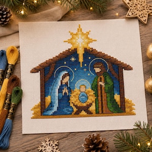Può includere: Un motivo a punto croce su tessuto bianco che raffigura una scena della natività con una stella gialla brillante sopra una stalla. Il design presenta figure in vesti blu e verdi che circondano un neonato centrale, ricamato con filo.