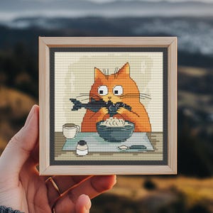 Peut inclure: Broderie au point de croix dans un cadre en bois. L'œuvre représente un chat orange mangeant un poisson dans un bol. Une tasse, un petit récipient et une cuillère sont sur la table. L'arrière-plan est flou.