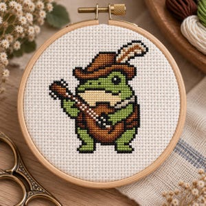 May include: A circular wooden embroidery hoop containing a cross-stitch pattern on white Aida fabric. The design features a green frog wearing a brown hat with a feather, playing a brown acoustic guitar, rendered in pixelated embroidery stitches.