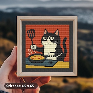 May include: Cross-stitch art in a wooden frame. The artwork depicts a black and white cat holding a spatula over a pan. The background is orange, with a yellow and blue base. The text "Stitches: 65 x 65" is at the bottom.