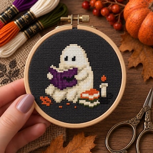 May include: A circular wooden embroidery hoop containing a cross-stitch design on black fabric. The design features a white ghost reading a purple book, with a stack of books, a lit candle, and scattered orange leaves on a dark background.
