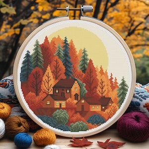 Op de afbeelding: Een kruissteekborduurwerk in een houten ring toont een herfstlandschap met huizen tussen bomen. Het kleurenpalet omvat tinten oranje, rood, groen en geel, met een grote gele zon op de achtergrond. De borduursel is omgeven door garen en herfstbladeren.