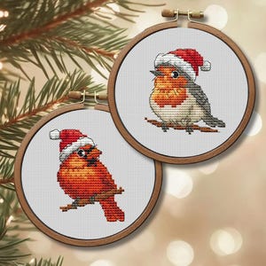 Pode incluir: Dois bastidores de ponto cruz apresentam pássaros festivos. Um cardeal vermelho e um pisco-de-peito-ruivo, ambos usando gorros de Papai Noel, estão empoleirados em galhos marrons. Perfeito para decorações de Natal.