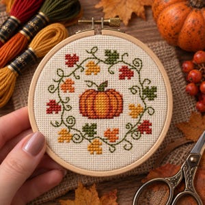 May include: A circular wooden embroidery hoop containing a cross-stitch design of a central orange pumpkin surrounded by a wreath of small red, yellow, and green autumn leaves and swirling vines on a light beige fabric background.