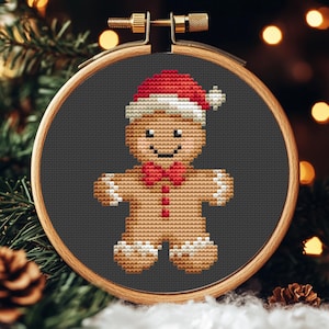 Könnte beinhalten: Kreuzstich-Stickerei eines Lebkuchenmanns mit Weihnachtsmütze und Fliege, in einem hölzernen Stickrahmen. Der Lebkuchenmann ist hellbraun mit weißen Akzenten, einem roten Hut und einer Fliege sowie einem schwarzen Lächeln. Der Hintergrund ist dunkelgrau.