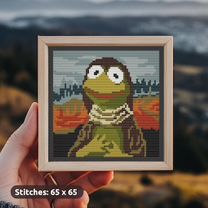 Op de afbeelding: Kruissteekkunst van een stripfiguur met grote ogen, een groen lichaam en een bruine kop, met een sjaal. Het kunstwerk is ingelijst in een licht houten frame met een donkergrijze rand. Tekst: Stitches: 65 x 65.