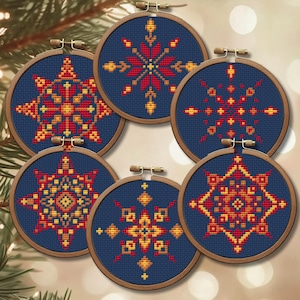 May include: Six round cross-stitch ornaments with wooden hoops. Each features a different geometric snowflake design in red, yellow, and orange on a navy blue background. The ornaments are arranged in a circular pattern.