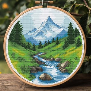 Könnte beinhalten: Ein Kreuzstich-Stickrahmen mit einer Berglandschaft. Das Design zeigt einen schneebedeckten Berg, einen Fluss, grüne Bäume und grasbewachsene Hügel. Der Rahmen ist aus hellem Holz gefertigt und vor einem natürlichen Hintergrund platziert.