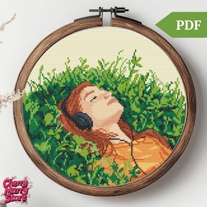 Op de afbeelding: Borduurpatroon van een vrouw met rood haar die in een veld met groen gras ligt, een koptelefoon draagt en naar muziek luistert.