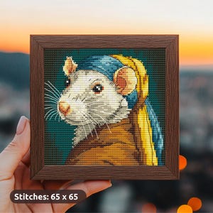 Könnte beinhalten: Kreuzstich-Kunstwerk einer Ratte mit blau-gelber Kopfbedeckung, gerahmt in einem braunen Holzquadrat. Die Ratte hat weißes Fell, eine rosa Nase und lange Schnurrhaare. Der Text "Stitches: 65 x 65" steht unten.