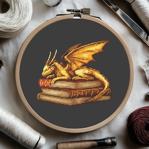 May include: Cross-stitch embroidery of a golden dragon resting on a stack of books. The dragon has detailed wings and is set against a dark gray background within a wooden embroidery hoop. The books are brown with gold lettering.