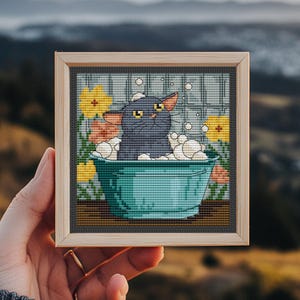 Pode incluir: Arte em ponto cruz de um gato preto tomando banho em uma banheira turquesa cheia de bolhas. O gato tem olhos amarelos e orelhas laranja. O fundo inclui flores amarelas e um padrão de grade cinza. A obra de arte é emoldurada em uma moldura de madeira marrom claro.
