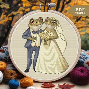 Può includere: Uno schema di punto croce di una coppia di rane che si sposano. Lo sposo indossa un abito blu e la sposa indossa un abito bianco con un velo. Lo schema è in un telaio da ricamo in legno.