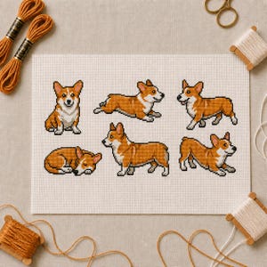 Può includere: Un motivo a punto croce su tessuto Aida bianco che raffigura sei cani Corgi in stile pixel art in varie pose, tra cui seduti, in corsa, in piedi e addormentati, realizzati con fili arancioni, bianchi e neri su una griglia.