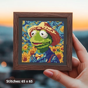 Könnte beinhalten: Ein gerahmtes Kreuzstichbild einer grünen Frosch, die einen braunen Hut trägt, umgeben von Sonnenblumen. Der Hintergrund ist eine blaue und gelbe Sternenhimmel-Szene. Das Bild ist 65 Stiche mal 65 Stiche groß.