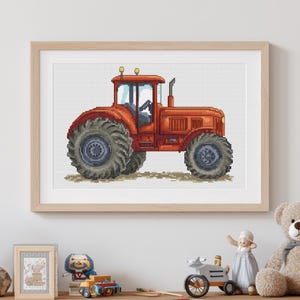 Peut inclure: Broderie au point de croix d'un tracteur rouge avec de grands pneus gris, sur fond blanc. Le tracteur est encadré dans un cadre en bois clair. L'œuvre est exposée sur une étagère avec d'autres jouets.