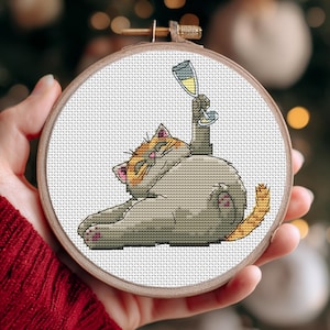 May include: A cross-stitch embroidery hoop featuring a cartoon cat holding a champagne glass. The cat is gray with orange and pink accents. The wooden hoop is held in a person's hand.