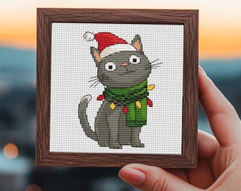 May include: A framed cross-stitch of a gray cat wearing a Santa hat and a green scarf adorned with Christmas lights. The cat has a smiling expression and is set against a white background. The frame is a dark brown color.