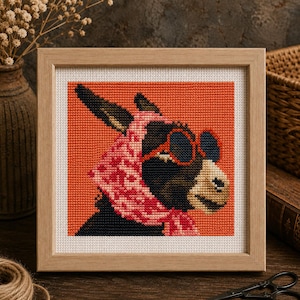 May include: A square cross-stitch embroidery in a light wood frame featuring a dark-colored donkey wearing a pink patterned headscarf and red-rimmed sunglasses against a solid orange background. The donkey has a tan muzzle and dark ears.