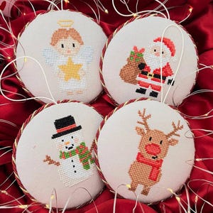 May include: Four round Christmas ornaments with cross-stitched designs. The ornaments feature an angel, Santa Claus, a snowman, and a reindeer. Each ornament has a cream-colored background, a red and gold rope border, and a hanging loop.