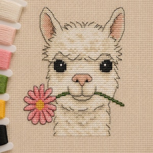 May include: A cross-stitch embroidery pattern on beige Aida fabric featuring a cream-colored llama with large black eyes and a pink nose, holding a pink daisy with a green stem in its mouth, accompanied by a vertical row of colorful embroidery floss.
