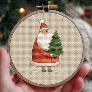May include: A cross-stitch embroidery hoop featuring a cartoon Santa Claus holding a green Christmas tree. Santa is wearing a red suit and hat with a white beard. The embroidery is set in a wooden hoop.