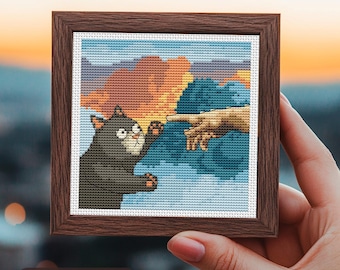 May include: A framed cross-stitch depicting a cat's version of Michelangelo's 'The Creation of Adam'. The cat, in shades of gray, reaches towards a human hand. The background features a sunset sky. The frame is a dark brown color. The text 'Stitches: 65 x 65' is visible.
