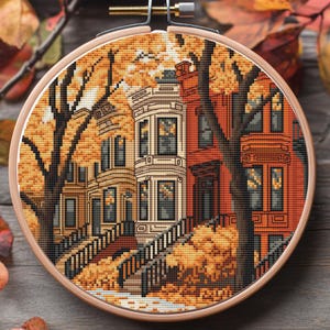 May include: A cross-stitch embroidery depicting a row of houses with a tree in front. The houses are painted in shades of red, white, and brown. The tree has yellow leaves and is set against a background of yellow leaves.