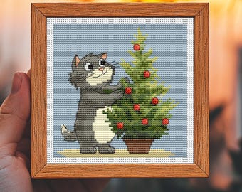 Könnte beinhalten: Ein gerahmtes Kreuzstichbild mit einer grauen Katze, die einen kleinen Weihnachtsbaum mit roten Kugeln schmückt. Die Katze steht auf ihren Hinterbeinen und greift nach dem Baum. Der Rahmen ist aus Holz.