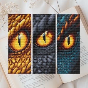 Op de afbeelding: Drie kruissteekpanelen met drakenogen in goud, zwart en blauwgroen. Elk oog heeft een gele iris en een donkere, verticale pupil. De panelen zijn geplaatst tegen de achtergrond van een open boek.