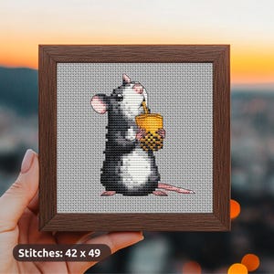 Puede incluir: Arte de punto de cruz en un marco marrón. La obra de arte representa una rata en blanco y negro sosteniendo una bebida amarilla con una pajita. El fondo es de color gris claro. El texto "Stitches: 42 x 49" está en la esquina inferior izquierda.
