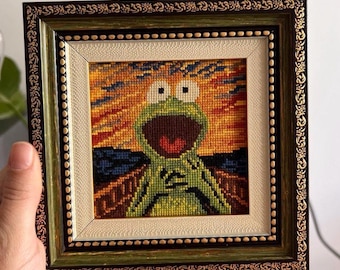 May include: A square cross-stitch embroidery featuring a green frog with a wide-open mouth, set against a vibrant orange, yellow, and blue background. The artwork is encased in a dark brown, ornate wooden frame with gold-toned detailing.