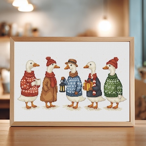 May include: Cross-stitch artwork in a wooden frame featuring five anthropomorphic geese. Each goose is dressed in winter attire, including sweaters, hats, and scarves. One goose holds a mug, another a lantern, and another a wrapped gift. The artwork is displayed on a wooden surface.