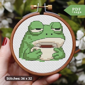 May include: Cross stitch pattern of a green frog holding a white mug with brown liquid. The frog has a tired expression and is sitting on a white background. The pattern is 36 stitches wide by 32 stitches high.