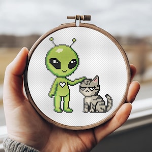 Puede incluir: Un bastidor de bordado de punto de cruz con un extraterrestre verde con un corazón, acariciando a un gato gris. La obra de arte está enmarcada en un círculo de madera, sobre un fondo blanco.