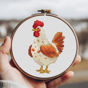 May include: A cross-stitch embroidery of a rooster in a wooden hoop. The rooster features a red comb and wattle, with white and orange feathers. The embroidery is on white fabric with a grid pattern.