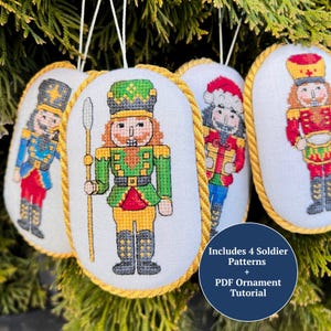 Puede incluir: Cuatro adornos navideños bordados con diseños de soldados cascanueces. Cada adorno tiene un fondo blanco y un borde de cuerda dorada. Los cascanueces visten uniformes variados con detalles en rojo, verde, azul y dorado. Los adornos cuelgan sobre un fondo verde y frondoso. Incluye 4 patrones de soldados + Tutorial de adorno PDF.
