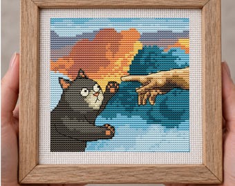 Pode incluir: Um bordado em ponto cruz quadrado numa moldura de madeira clara, apresentando um gato cinzento a estender a pata para tocar num dedo humano. O fundo retrata um pôr do sol colorido com tons de laranja e azul, imitando uma composição clássica.