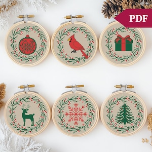 May include: Six embroidery hoop ornaments with a Christmas theme. Each hoop features a different design: a red bauble, a cardinal, a gift box, a reindeer, a snowflake, and a Christmas tree. All designs are surrounded by a green wreath with red berries.