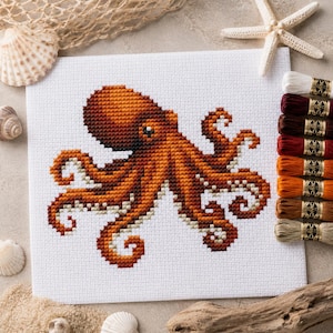 Peut inclure: Un motif de point de croix sur tissu blanc représentant une pieuvre brune aux huit tentacules enroulés. Le design est entouré de coquillages, d'une étoile de mer et d'un filet, avec une pile verticale de huit échevettes de fil à broder.